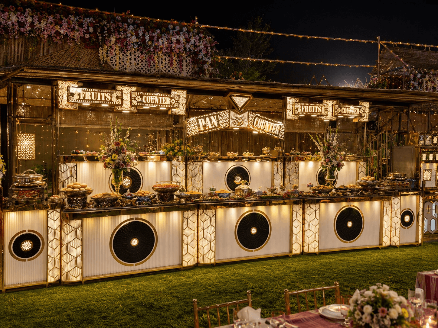 Illuminated fruit and pan buffet counters on a lawn at night